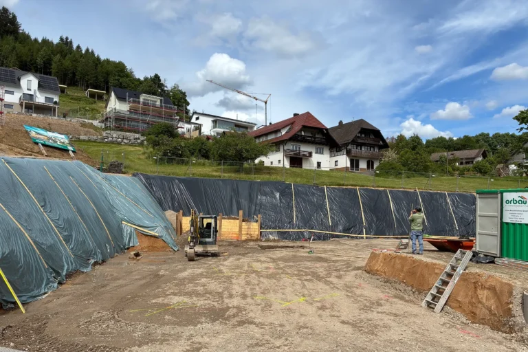 Große Baugrube für das Mehrgenerationenhaus in Fischerbach mit gesicherten Böschungen, Minibagger und markierten Leitungsverläufen. Die Baustelle liegt eingebettet in die Schwarzwaldlandschaft.
