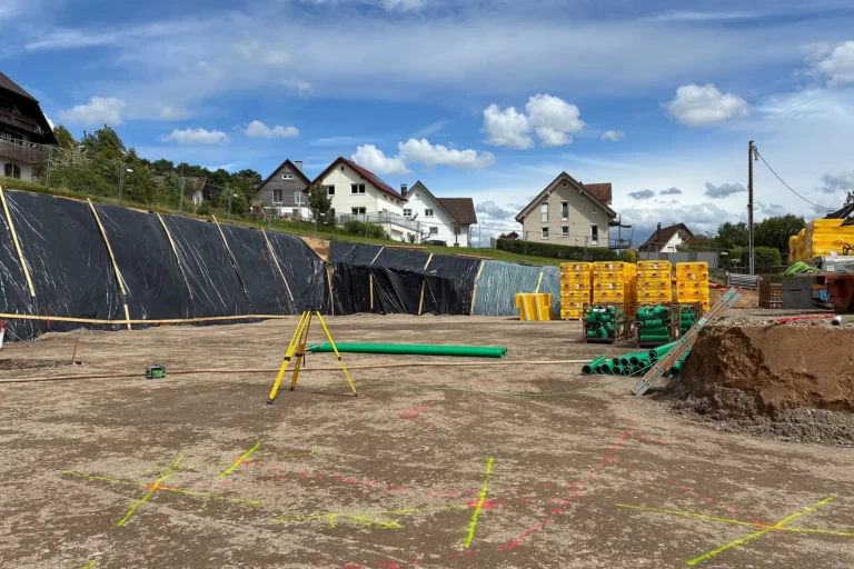 Baugrube des Mehrgenerationenhauses in Fischerbach mit vorbereiteter Bodenplatte, Vermessungsgerät, gelagerten Baumaterialien und gesicherter Böschung. Im Hintergrund sind Wohnhäuser und der Schwarzwaldhimmel zu sehen.