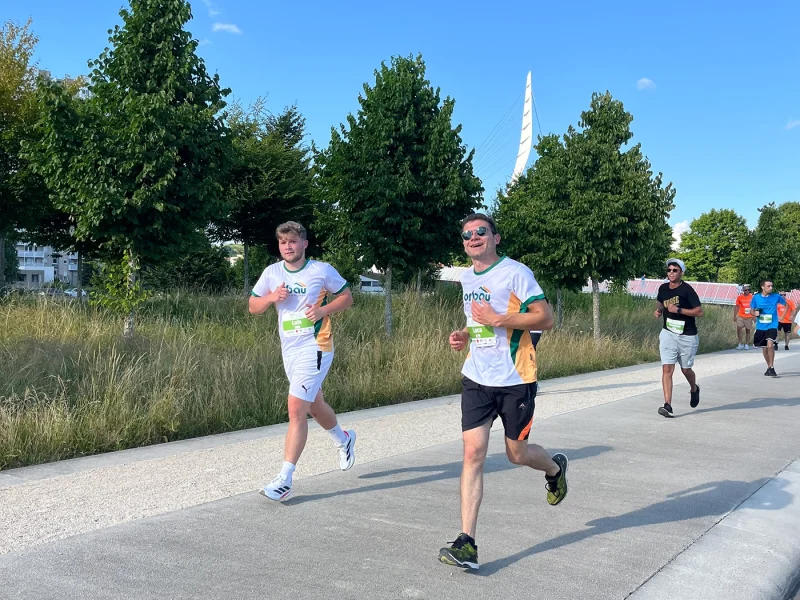 Zwei orbau-Läufer joggen nebeneinander auf der Laufstrecke, Bäume im Hintergrund.
