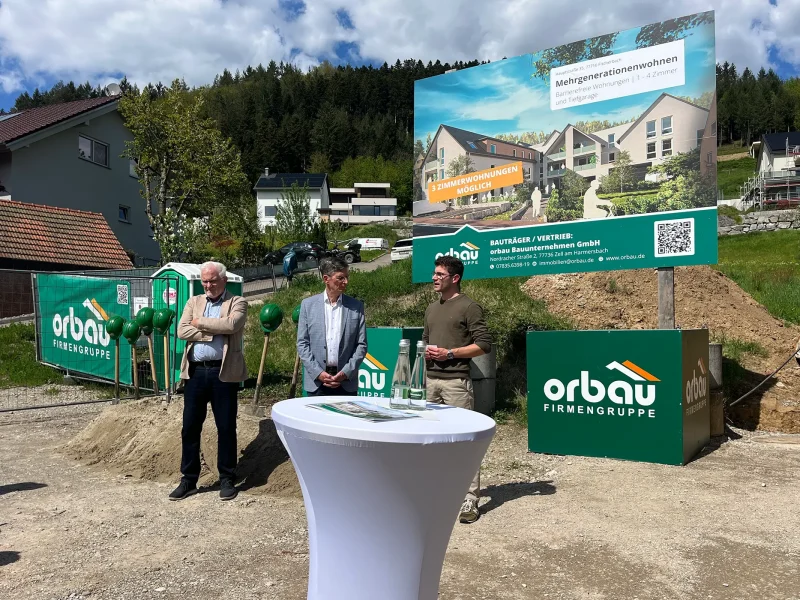 Ansprache beim Spatenstich in Fischerbach mit Projektverantwortlichen vor Baustelle und orbau Banner