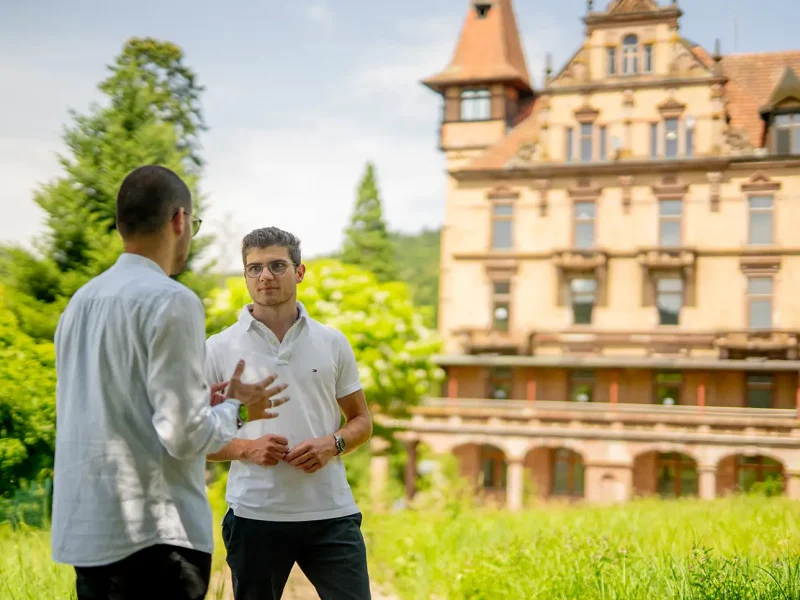 Die beiden Architekten des Schloss Rothschild stehen vor dem denkmalgeschützen Gebäude bei strahlendem Sonnenschein.